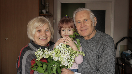 Happy granddaughter embraces senior smiling grandfather with grandmother. Hugs. Grandparents couple with their kid at home. Senior lifestyle family concept. Family time relax with young child girl