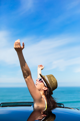 Joyful woman on summer travel vacation to the coast  leaning out car sunroof towards the sea.