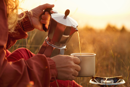 Hiker woman pours hot coffee into cup with coffee pot on background of sunrise meadow in mountains. Tourist makes transition, meets dawn sun. Lens flare. Campaign. Life style. Travel