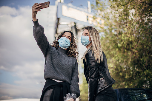 Two young women wearing protective masks and making selfie with smart phone on the city street.