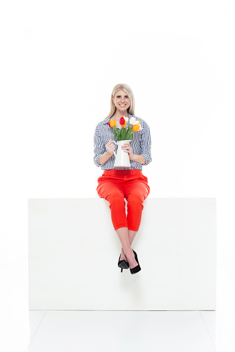 One person of aged 20-29 years old who is beautiful with blond hair caucasian female businesswoman sitting in front of white background wearing business casual and high heels who is showing cool attitude and holding tulip