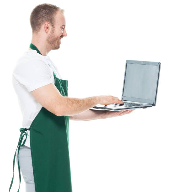 istock Caucasian young male standing in front of white background wearing apron and using computer 1223649327