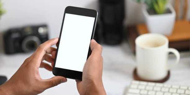 istock Cropped image of hands holding a cropped black smartphone with white blank screen over working desk that surrounded by office equipment as background. 1223654496