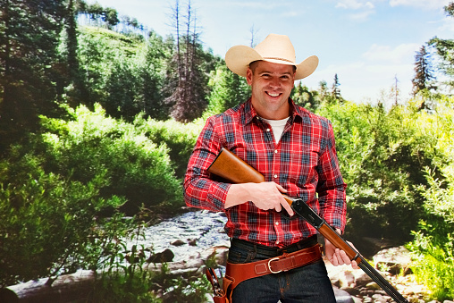 Waist up of with short hair caucasian young male cowboy in front of landscape - scenery who is outdoors wearing cowboy hat who is smiling who is shooting a weapon and holding weapon and using rifle