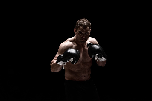 Front view of with brown hair caucasian male in front of black background wearing shorts who is confident and boxing and showing fist who is fighting