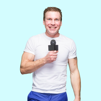 Front view of with brown hair caucasian young male sportscaster standing in front of colored background wearing pants who is talking and holding microphone