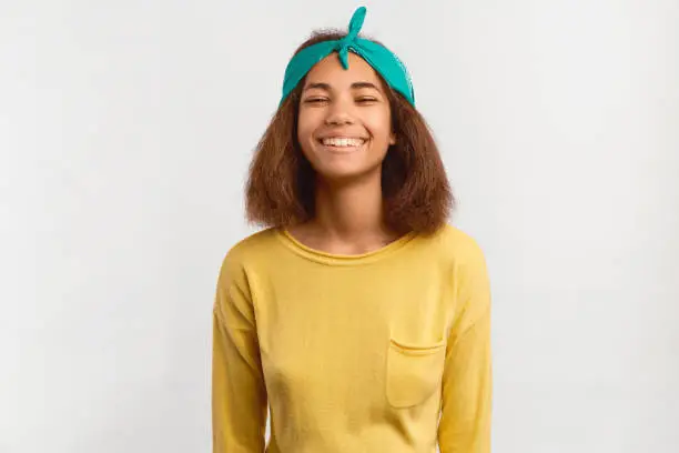 Photo of Girl feels ecstatic, laughs, lifts chin, looks at camera with half-closed eyes. Happy African-American teenage girl celebrates victory, wearing green bandana and yellow sweater isolated on white wall