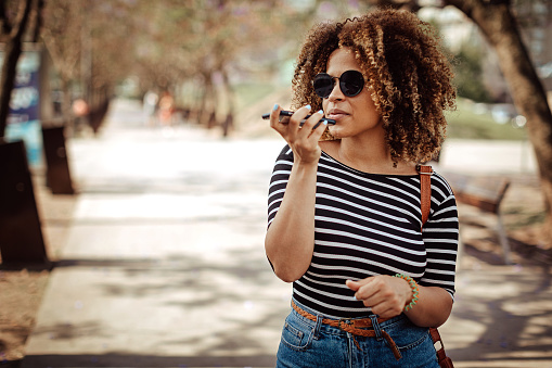 Young Latin woman with big curly hair and sunglasses on sending voice message on smart phone on the street