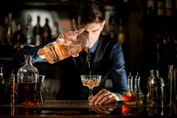 Barman holding glass of cocktail and pours it into wineglass. Young bartender in blue shirt holding glass of cold alcoholic cocktail and pours it into wineglass. bartender photos stock pictures, royalty-free photos & images