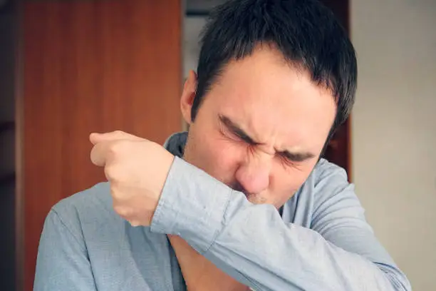 man having the flu and sneezing on her sleeve in the crook of her arm. A young guy coughs into the fabric of his shirt to prevent the spread of viral bacteria and not infect other people .