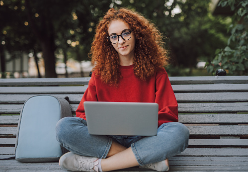 Positive young woman with laptop sitting on bench in city