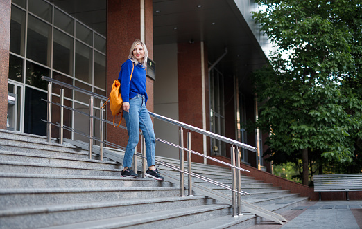 Full body young woman with backpack smiling and looking away while walking down steps after university lessons