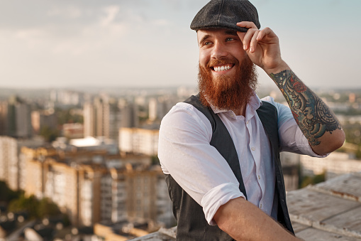 Bearded young man with tattooed arm smiling and adjusting trendy hat while sitting on border on roof of building on blurred background of modern city