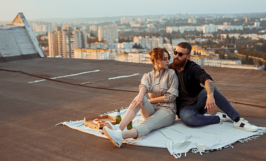 Young man and woman in stylish clothes sitting on blanket near beer and pizza during romantic date on rooftop in evening