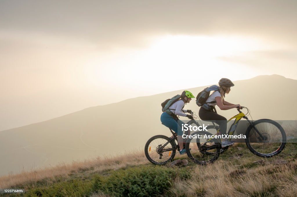 Two women ride up grassy hillside on electric mountain bikes Sun sets over distant mountain range Electric Bicycle Stock Photo