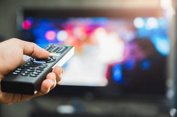 istock Young man holding television remote control. Hands pointing to tv screen set and turning it on or off select channel watching tv on his sofa at home in the living room relax. 1223732978