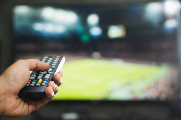 istock Young man holding television remote control watching football program. Hands pointing to tv screen set and turning it on or off select channel watching tv in the living room relax. 1223733138