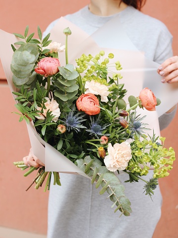 A woman in a grey dress holding a colorful fresh bouquet of flowers (pink carnations, fritillary, coral ranunculus, eucalyptus, eryngium) wrapped in light pink paper on the coral pink wall background.