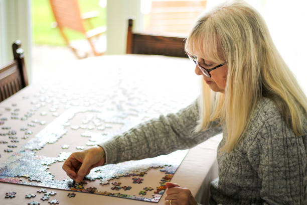 Color image depicting a senior caucasian couple, in their 60s and 70s, doing a jigsaw puzzle together at home during the coronavirus (Covid-19) pandemic. The couple are both wearing spectacles and are concentrating on completing the puzzle together. Room for copy space.