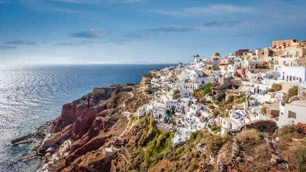 Panorama view towards Oia with the iconic white and blue typical greek Santorinia Houses, Windmill and orthodox church at the rocky mediterranean coast of Santorini. Oia, Santorini Island, Cyclades Islands, Greece, Europe.