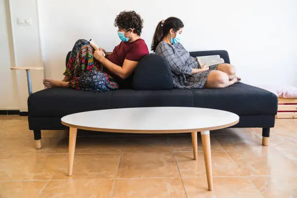 New normal situation after the 2020 coronavirus covid 19 sanitary crisis, a spanish couple rest on a black couch reading books while wearing a surgical mask.