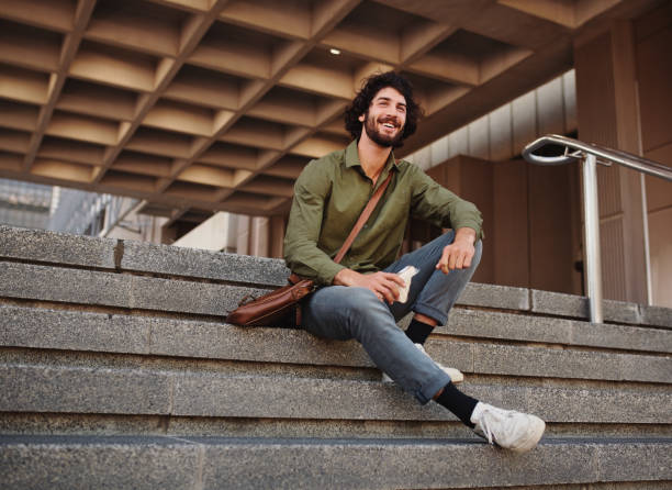Handsome young man eating sandwich while sitting on stairs during break while looking away