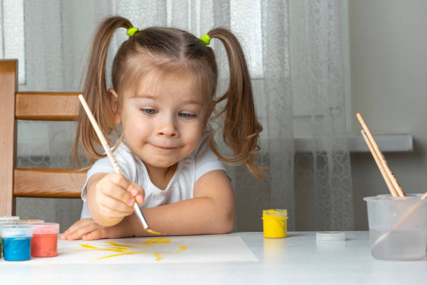 little beautiful girl 3-4 years old with two ponytails in a white t-shirt paints at the table and smiles. quarantined children's creativity due to coronavirus - child caucasian little girls 3 4 years imagens e fotografias de stock