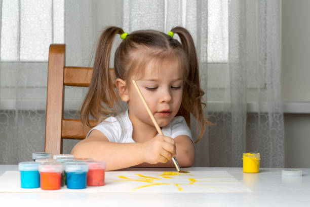 little beautiful girl 3-4 years old with two ponytails in a white t-shirt draws paints at the table. quarantined children's creativity due to coronavirus - child caucasian little girls 3 4 years imagens e fotografias de stock