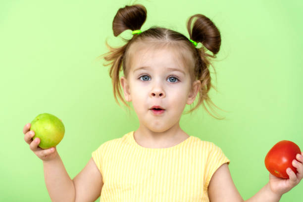 une petite fille de 3 à 5 ans dans un t-shirt jaune tient émotionnellement une pomme et une tomate dans ses mains. le problème du manque de fruits et légumes dans les aliments pour bébés - child caucasian little girls 3 4 years photos et images de collection