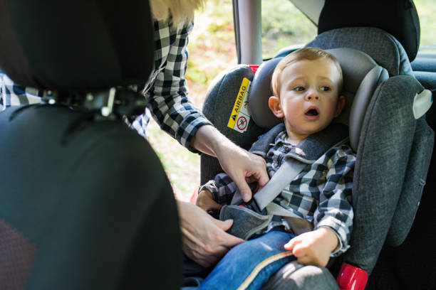 woman is fastening security belt to happy child, who is sitting in safety car seat stock photo