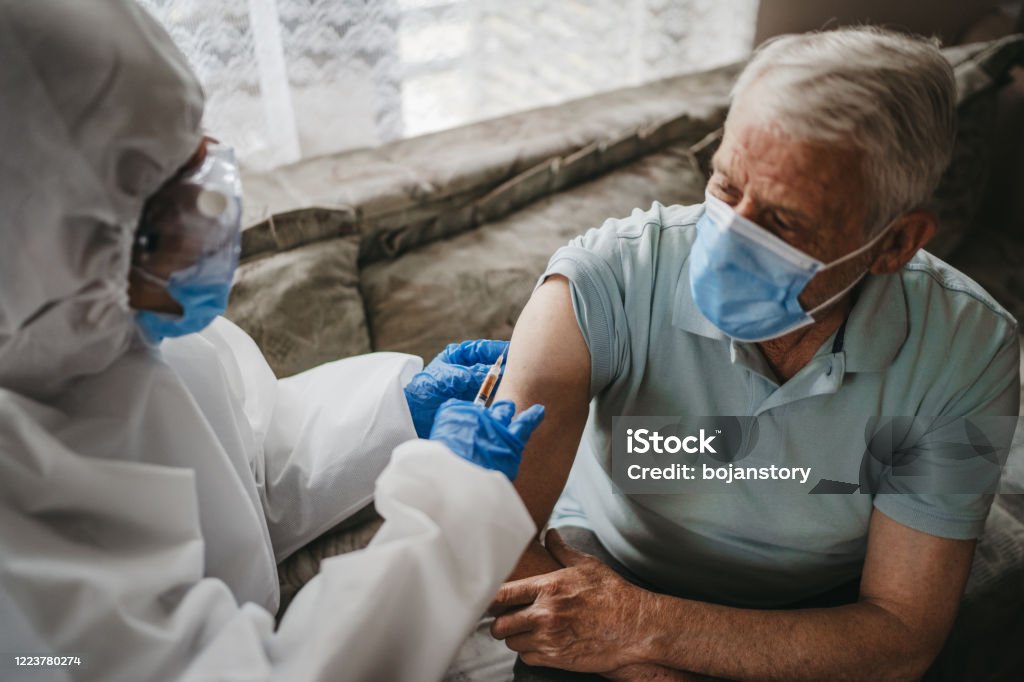 Injection Female doctor in protective suit giving vaccine against covid19 virus or injection to senior man at home. Therapy at home in pandemic conditions Vaccination Stock Photo