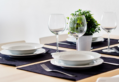 Close up new white porcelain tableware plates on purple place mats and empty wine glasses ready for dinner. Table settings wait for guests at home or restaurant, artificial potted plant for decoration