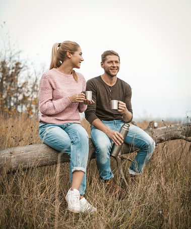 Smiling couple of tourists drink tea and talk while sitting on an old fallen tree among an autumn nature, pretty blonde and a Caucasian man in Casual are resting at sunset.