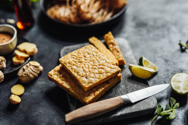 Preparing a vegan dish with tempeh Close-up of tempeh on cutting board with kitchen knife with other ingredients on table. Preparing a vegan dish in kitchen. meat substitute stock pictures, royalty-free photos & images