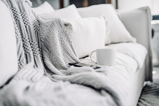 A coffee cup and smartphone on a white sofa, with cushions and a blanket