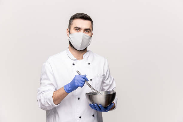 joven chef masculino en uniforme, máscara protectora y guantes mezclando leche y huevos crudos - scientist lab coat doctor male fotografías e imágenes de stock