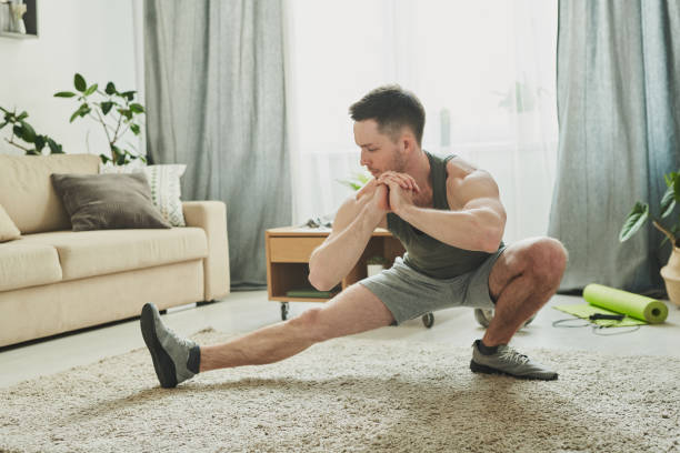 istock Young active man in sportswear doing stretching exercise for legs on the carpet 1223817485