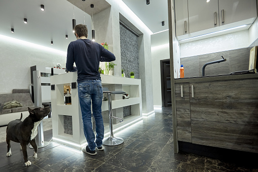 Young man cooking on modern kitchen. High tech interior in male style, gray concrete, dark wooden surfaces and black ceramic floor, designer light system.