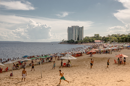 Group of local Brazilians play beach soccer while people relax sitting on chairs and swim in the black Rio Negra river at the urban Ponta Negra beach in Manaus located in the Amazon region of Brazil, South America