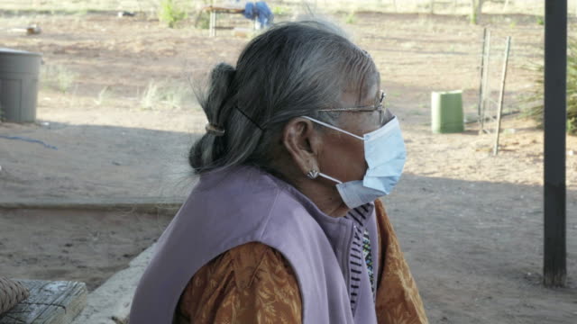 Navajo Grandma in her 80s sitting in front of her house in Monument Valley Arizona wearing a face mask to prevent contracting the Corona or Covid-19 Virus