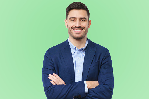 Handsome young man in formal blazer, standing in front of camera with positive confident smile, holding arms crossed, feeling relaxed, isolated on green background