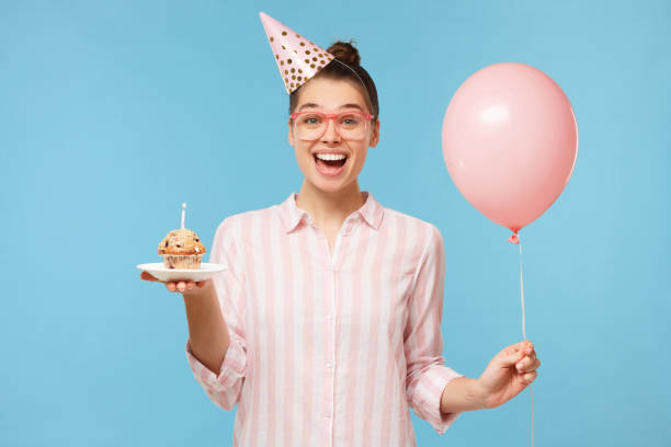 young excited girl in birthday hat, holding pink balloon in one hand and festive cake in another, laughing happily, isolated on blue background - personal accessory balloon beauty birthday imagens e fotografias de stock