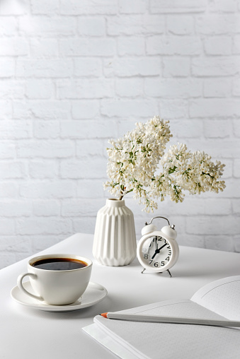 Cup of coffee, notebook, pencil, alarm clock and a vase with lilac flowers on a white table on a background of a white brick wall. Composition with white objects on a white background. Business morning concept with selective focus.