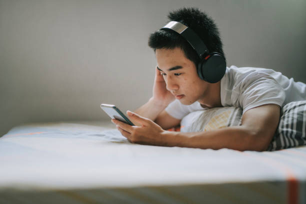 un ragazzo adolescente cinese asiatico a casa che ascolta musica con le cuffie durante il fine settimana sdraiato a letto - smart phone china mobile phone asia foto e immagini stock