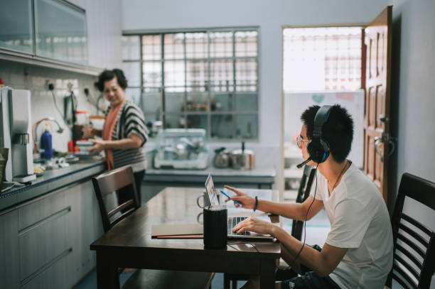 ein asiatischer chinesischer teenager, der online mit all seinen klassenkameraden über online-software plaudert, während er seine hausaufgaben am esstisch in der küche macht, während seine großmutter in der küche kocht und essen zubereitet - hausaufgabe fotos stock-fotos und bilder