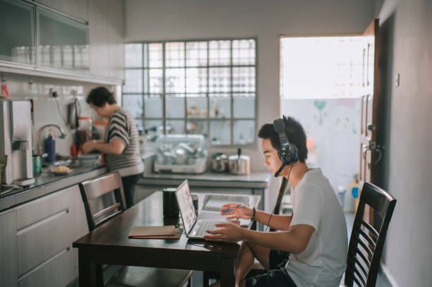 an asian chinese teenager chatting online with all his classmates via online software while doing his homework at the kitchen dining table while his grandmother cooking in the kitchen preparing food - grandparent using computer laptop dining table imagens e fotografias de stock