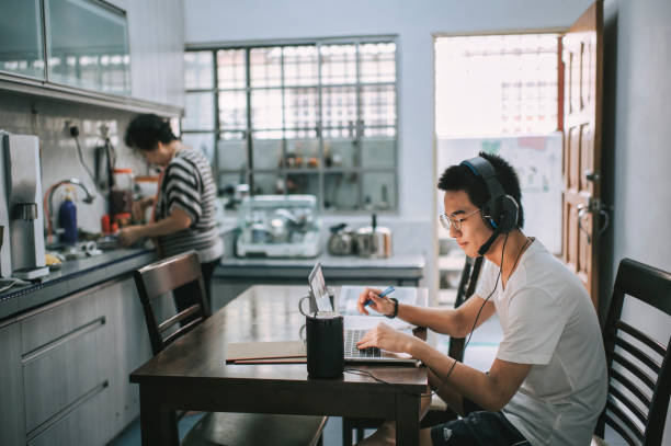 an asian chinese teenager chatting online with all his classmates via online software while doing his homework at the kitchen dining table while his grandmother cooking in the kitchen preparing food - grandparent using computer laptop dining table imagens e fotografias de stock