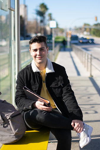 Young student guy sitting cross-legged at the bus stop holding a notebook while looking at the camera with a smile
