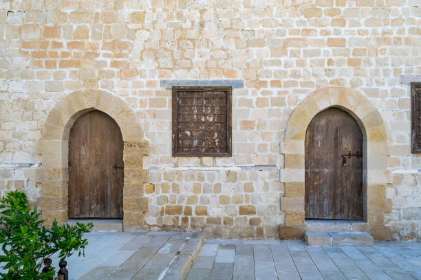 Two weathered wooden arched doors and one closed rusted wrought iron window on bricks stone wall and tiled stone floor