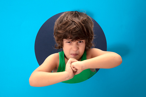 Happy child in a green t-shirt and hands poses through torn paper hole. Effect of blue torn paper.Child's face looking through a hole in blue pop art paper background.Boy peering through hole.